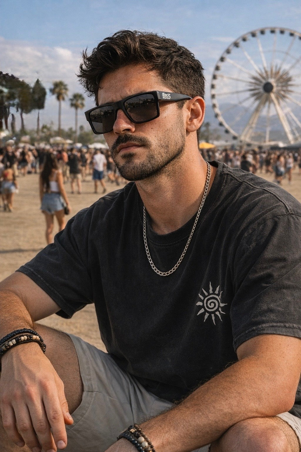 Man wearing square frame sunglasses and a black t-shirt with a sun logo, sitting outside at a festival.